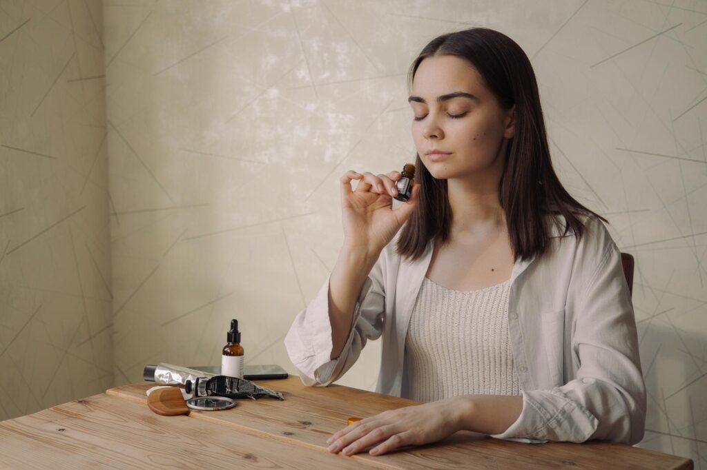 Mulher sentada à mesa, de olhos fechados, sentindo o aroma de um pequeno frasco de fragrância que segura perto do rosto